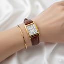 An elegant wrist shot of the Raymond Weil 5722 watch, featuring its rectangular gold case and brown leather strap, styled with a slender gold bangle on a white satin-like surface.