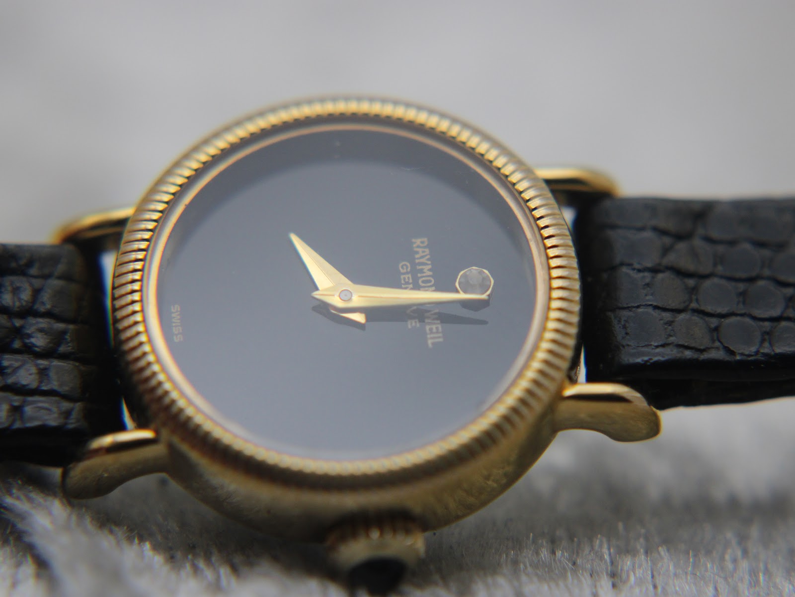 Detail shot of the watch dial, focused on the gold hands, Raymond Weil Genève text, and the diamond-style marker.
