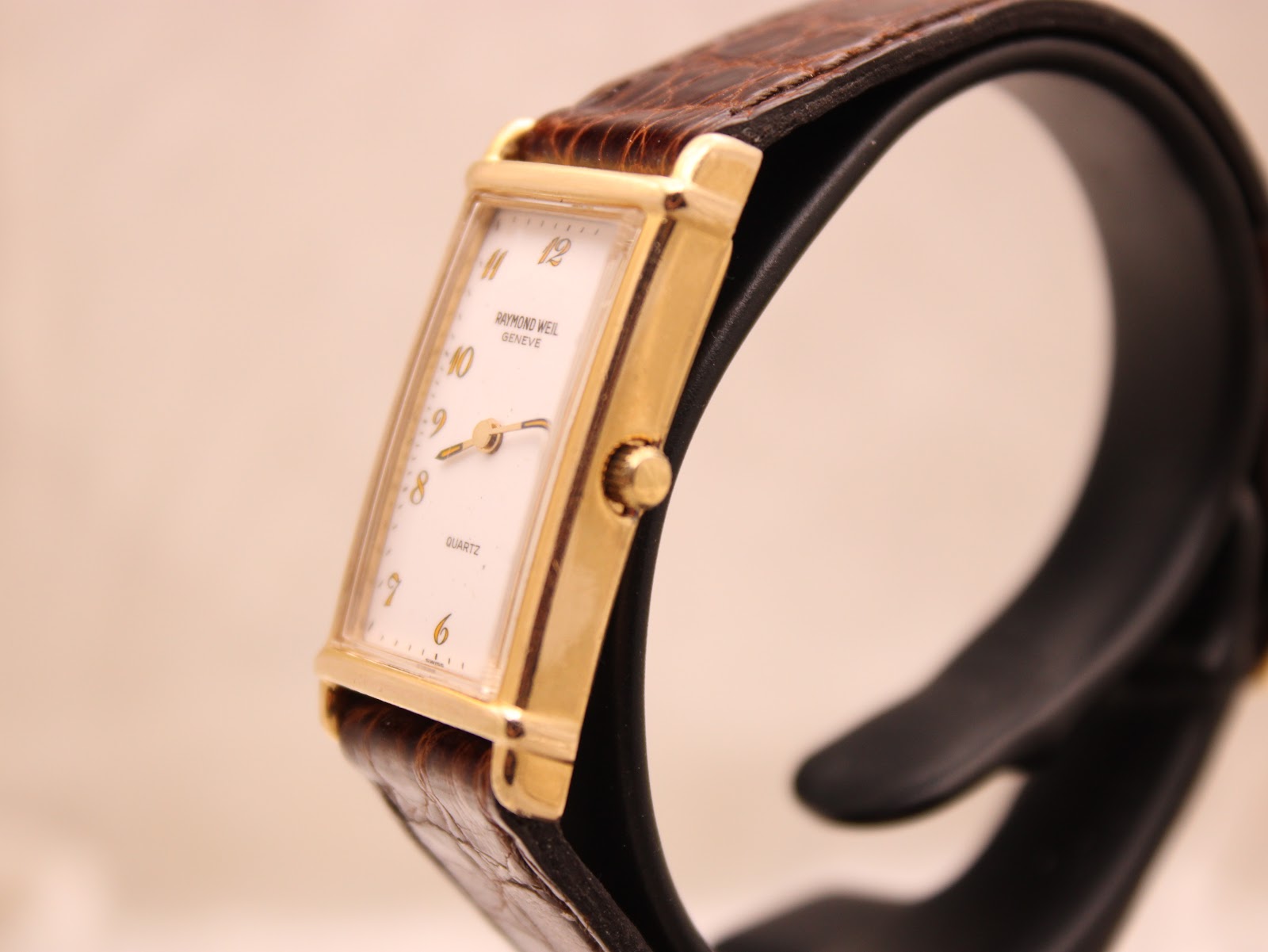 Side profile view of the Raymond Weil watch on a display stand, showing the gold case and crown