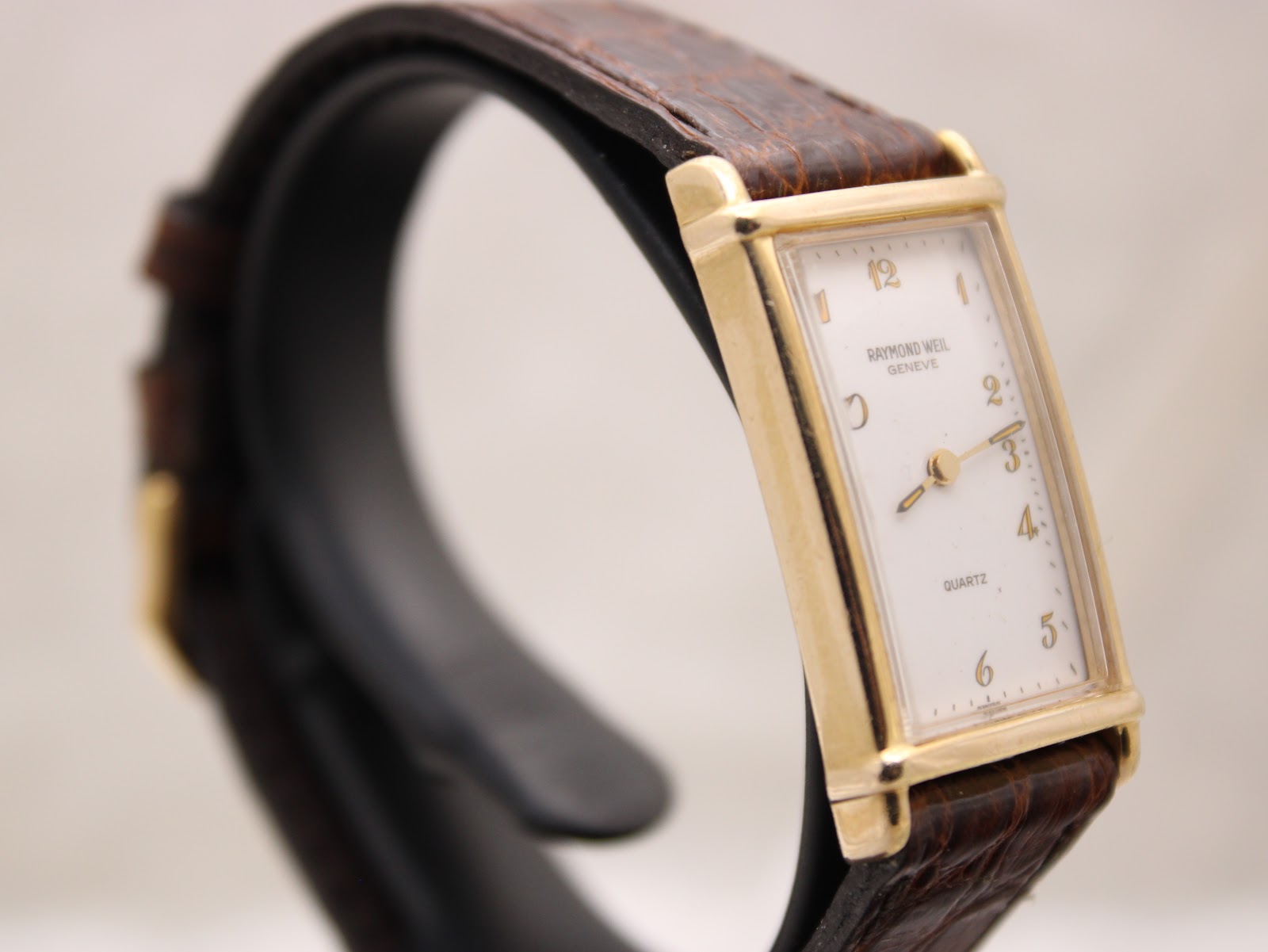 Three-quarter view of the Raymond Weil watch on a display stand, highlighting the rectangular gold case and brown strap