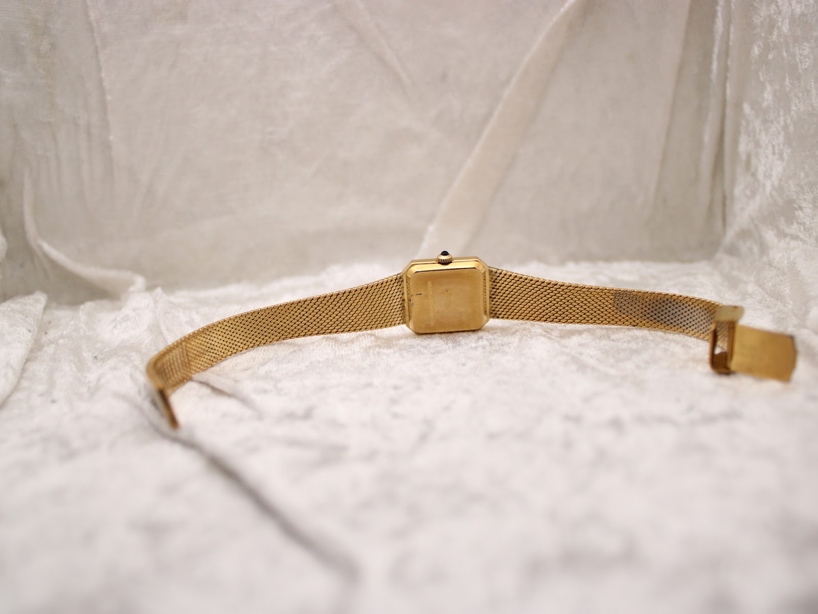 Rear view of the watch on a white textured surface, showing the plain case back and extended mesh bracelet.