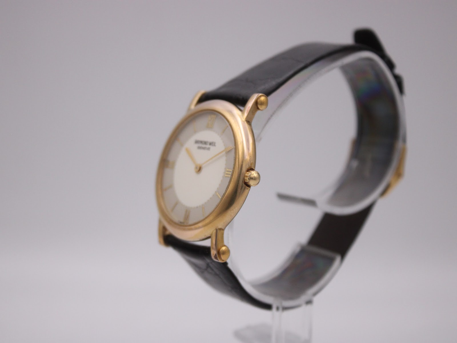 Angled right side view of the Raymond Weil watch showing the gold case, cream dial, and black leather strap on a clear display stand.