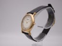 Angled right side view of the Raymond Weil watch showing the gold case, cream dial, and black leather strap on a clear display stand.