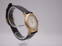 Angled left side view of the Raymond Weil watch, highlighting the gold-plated crown and case profile on a clear display stand.