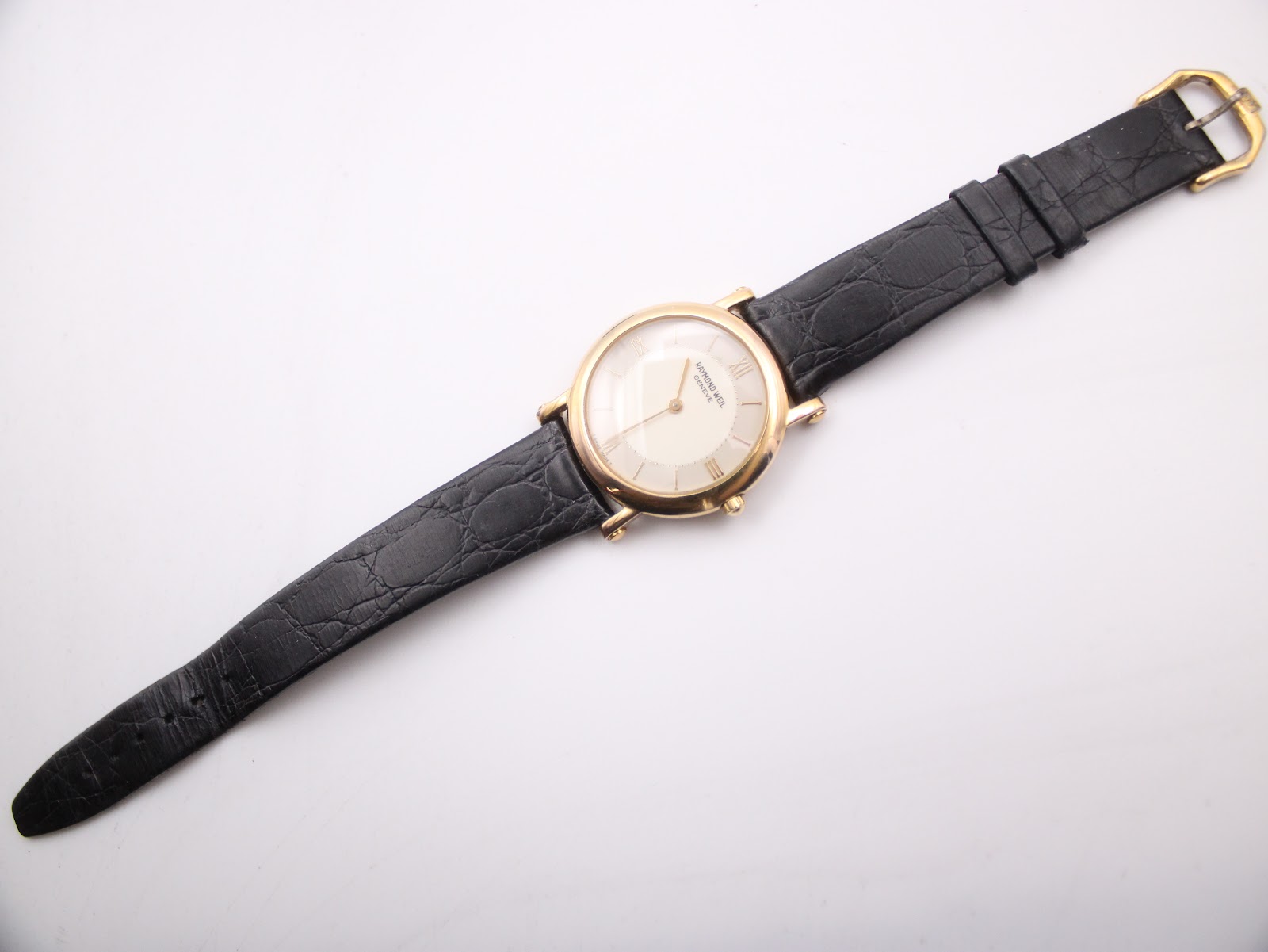 Overhead view of the entire Raymond Weil watch, strap extended, on a white surface, showcasing the dial and black leather strap.