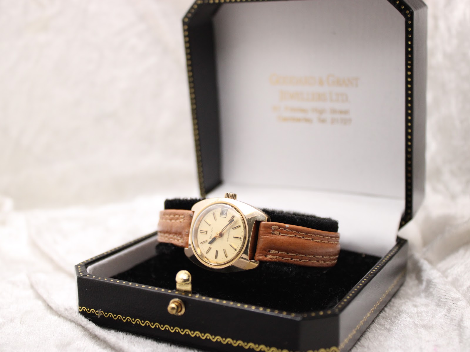 Bulova watch displayed in an open black jewellery box with a white interior, showing the dial and brown leather strap.