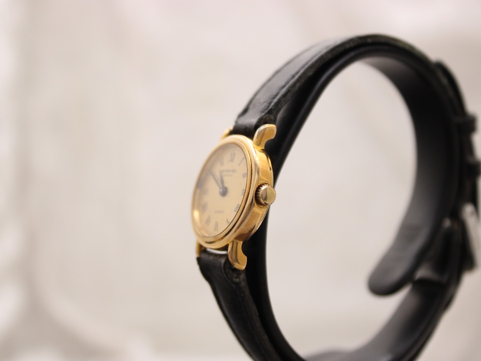 Side view of the Raymond Weil watch on a stand, showing the gold-tone crown and the curved profile of the case.