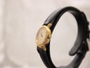 Side view of the Raymond Weil watch on a stand, showing the gold-tone crown and the curved profile of the case.