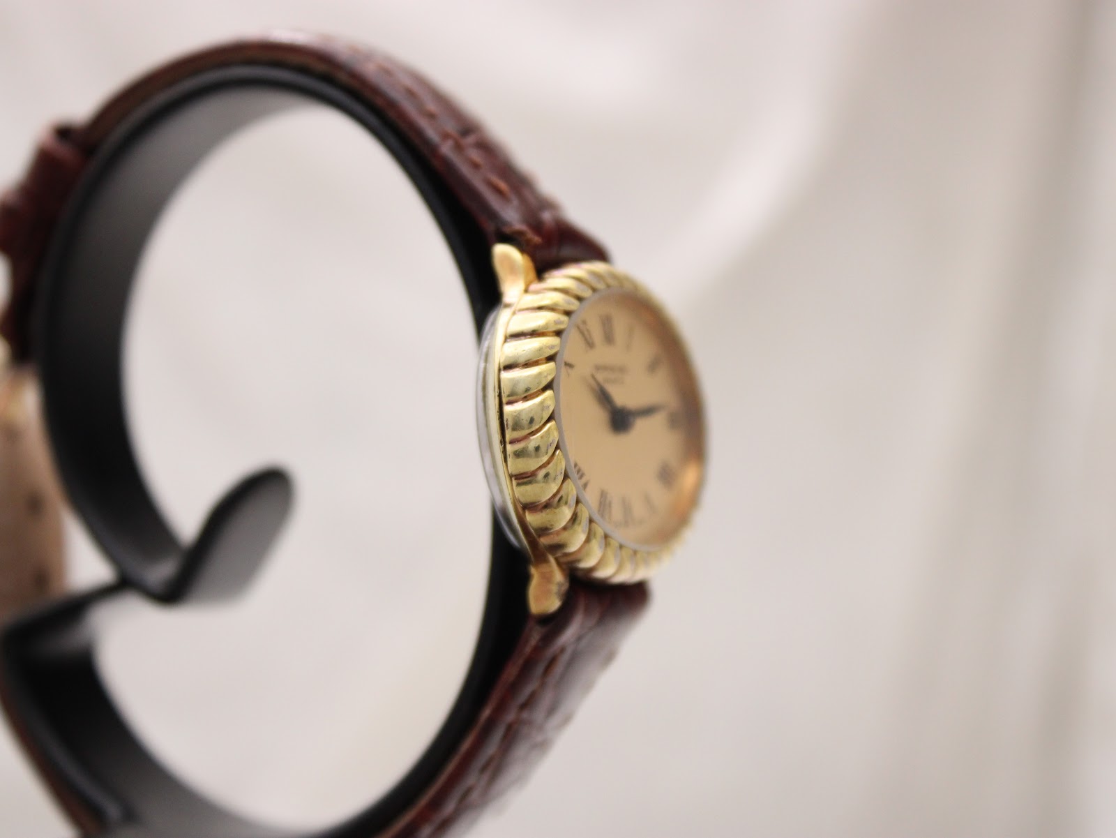 Angled side view of the Raymond Weil 683 watch on a stand, showing the curved gold-tone case and part of the brown leather strap.
