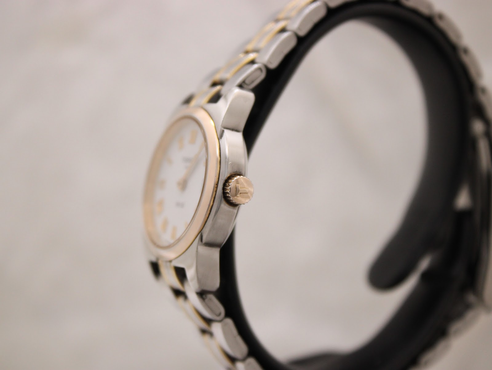 Side profile view of the Tissot PR 50 watch, showing the gold-tone crown and two-tone bracelet on a stand.