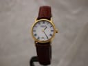Front view of Raymond Weil Genève 9923 gold electroplated watch with white Roman numeral dial and brown leather strap on a watch stand.