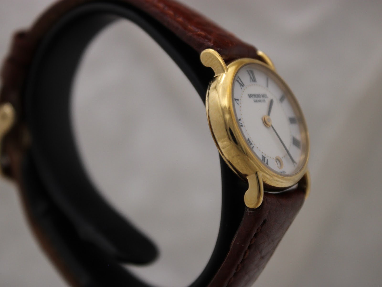 Left side view of the Raymond Weil 9923 watch, showing the polished gold electroplated case and brown leather strap.
