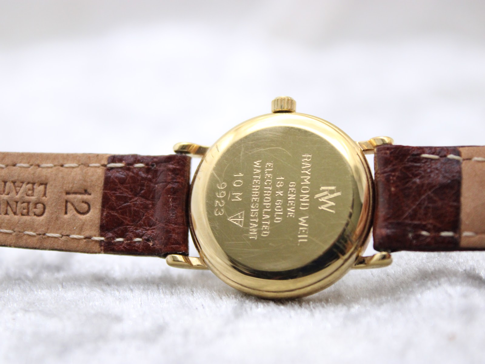 Close-up of the gold-tone case back of the Raymond Weil 9923, engraved with model number, water resistance, and "18K Gold Electroplated", and "12" on the strap end.