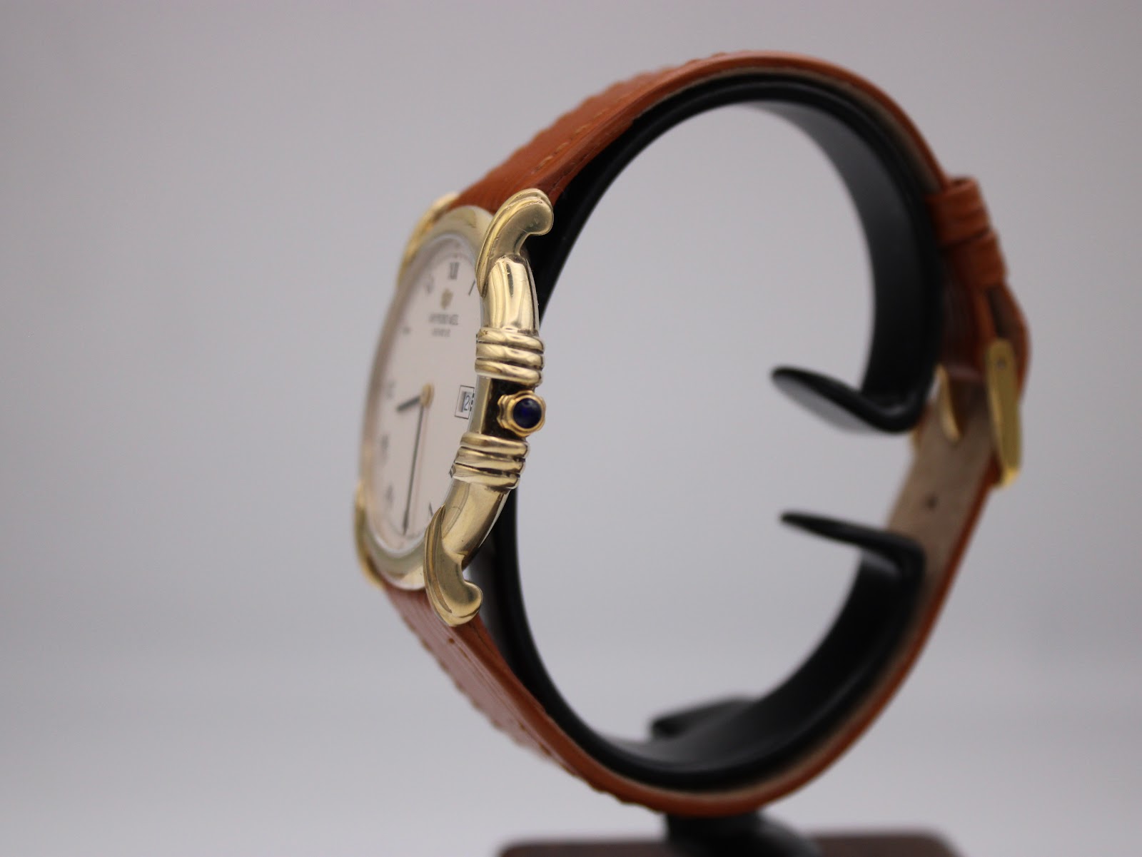 Side view of the Raymond Weil 9155 watch showing the gold-plated case, blue cabochon crown, and brown strap on a stand.
