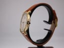 Side view of the Raymond Weil 9155 watch showing the gold-plated case, blue cabochon crown, and brown strap on a stand.
