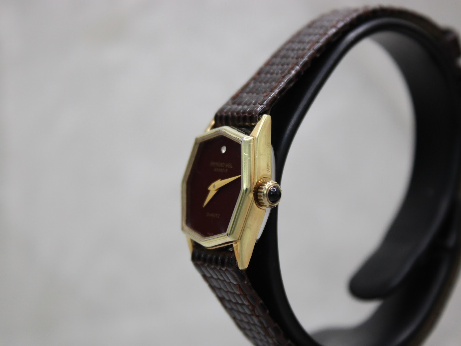 Side view of the watch on a stand, highlighting the gold-plated hexagonal case and black cabochon crown.
