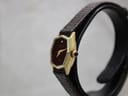 Side view of the watch on a stand, highlighting the gold-plated hexagonal case and black cabochon crown.
