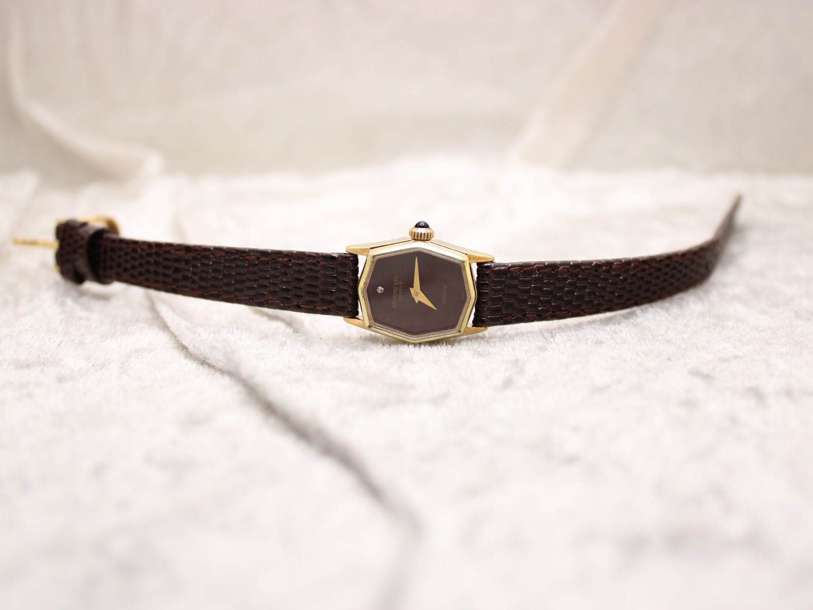 Overview of the watch laid flat on a textured white surface, showing the brown hexagonal dial and full length of the brown lizard-style strap.