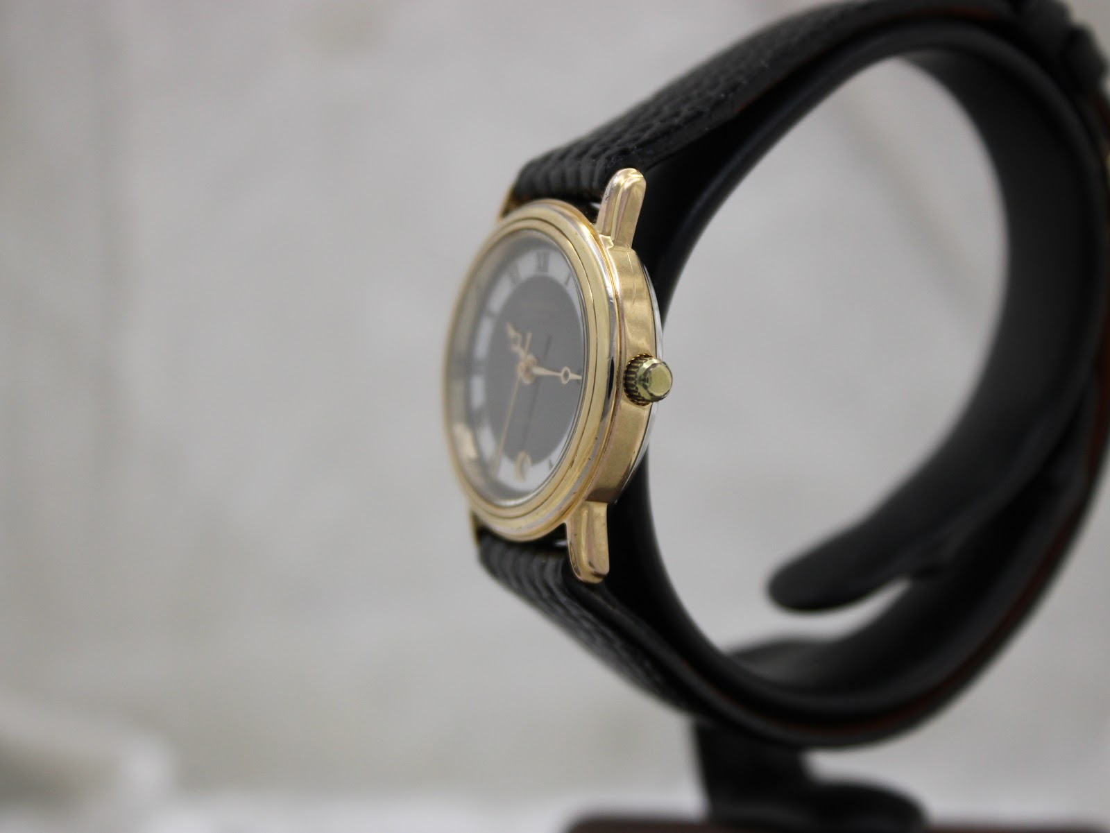 Side profile of the gold-tone Raymond Weil watch, showing the crown and polished case on a black watch stand.