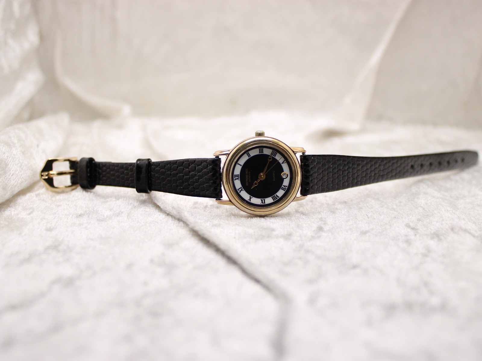 Full length view of the Raymond Weil watch with its black leather strap laid flat on a white textured surface, showing the gold-tone buckle.
