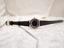 Full length view of the Raymond Weil watch with its black leather strap laid flat on a white textured surface, showing the gold-tone buckle.