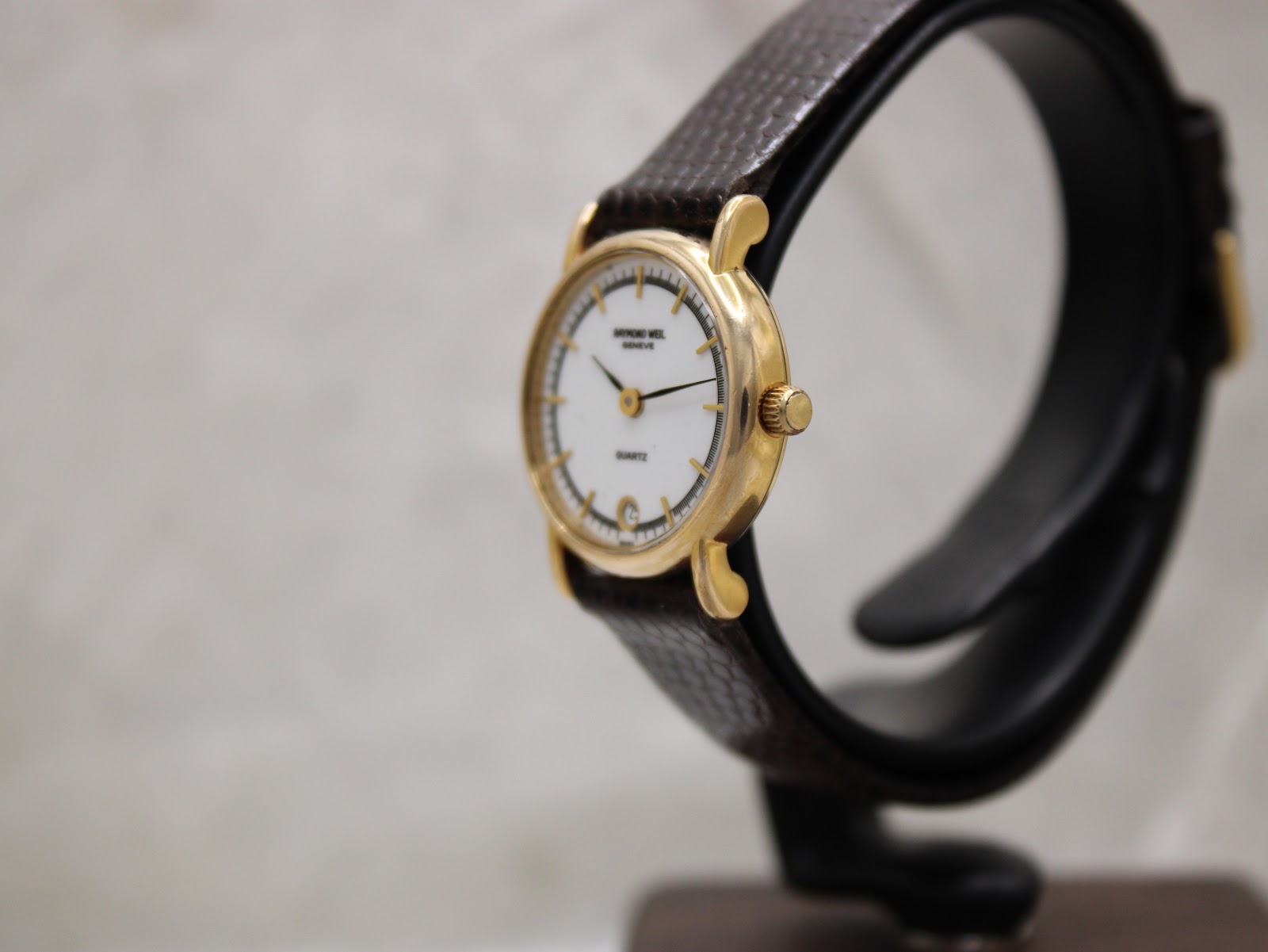 Angled side view of the Raymond Weil watch, showing the gold-tone case, crown, and texture of the brown leather strap.