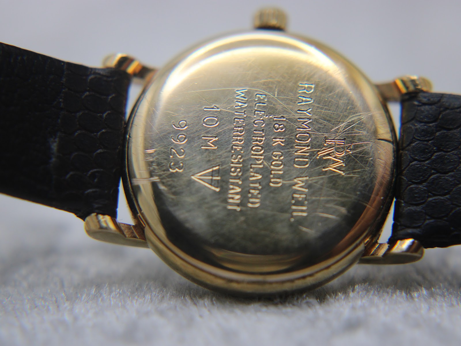 Close-up of the gold-tone case back of the Raymond Weil watch, showing engravings including 'RAYMOND WEIL GENEVE', '18K GOLD ELECTROPLATED', and '9923'.