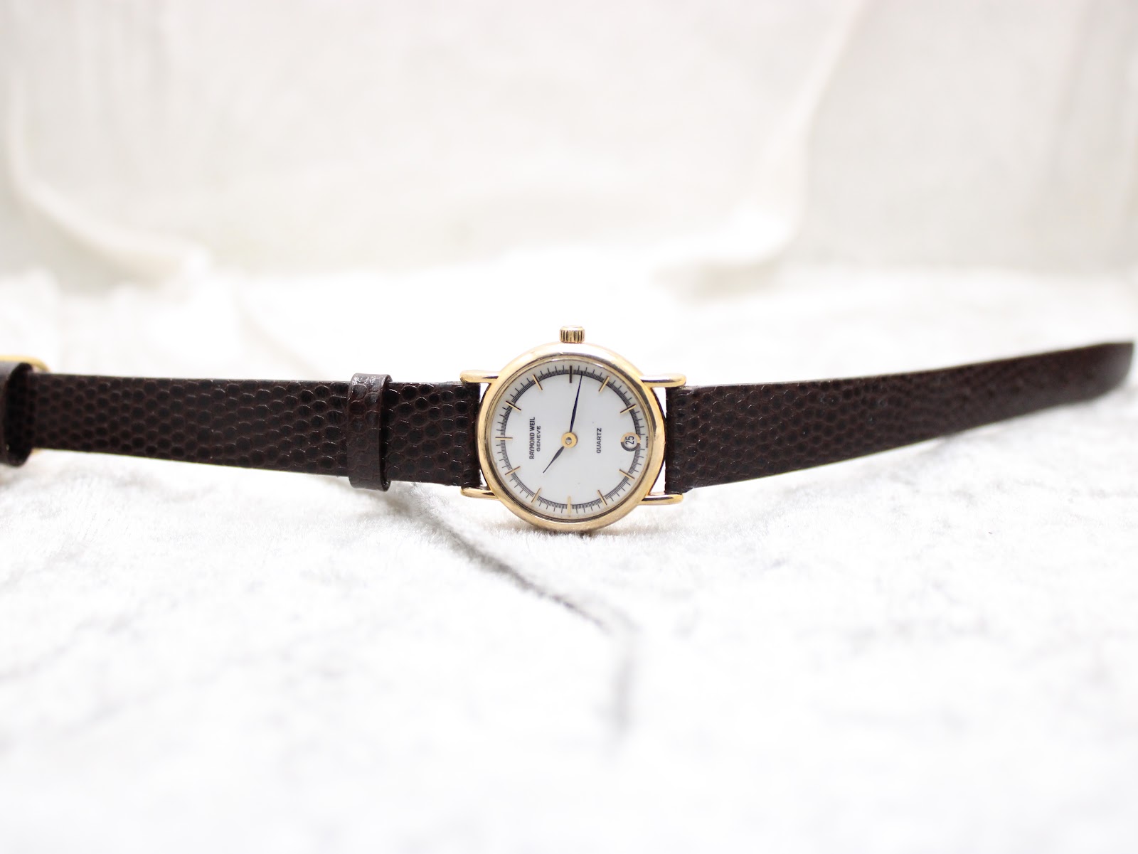 Top-down view of the Raymond Weil watch with its brown leather strap fully extended, lying flat on a white textured surface.