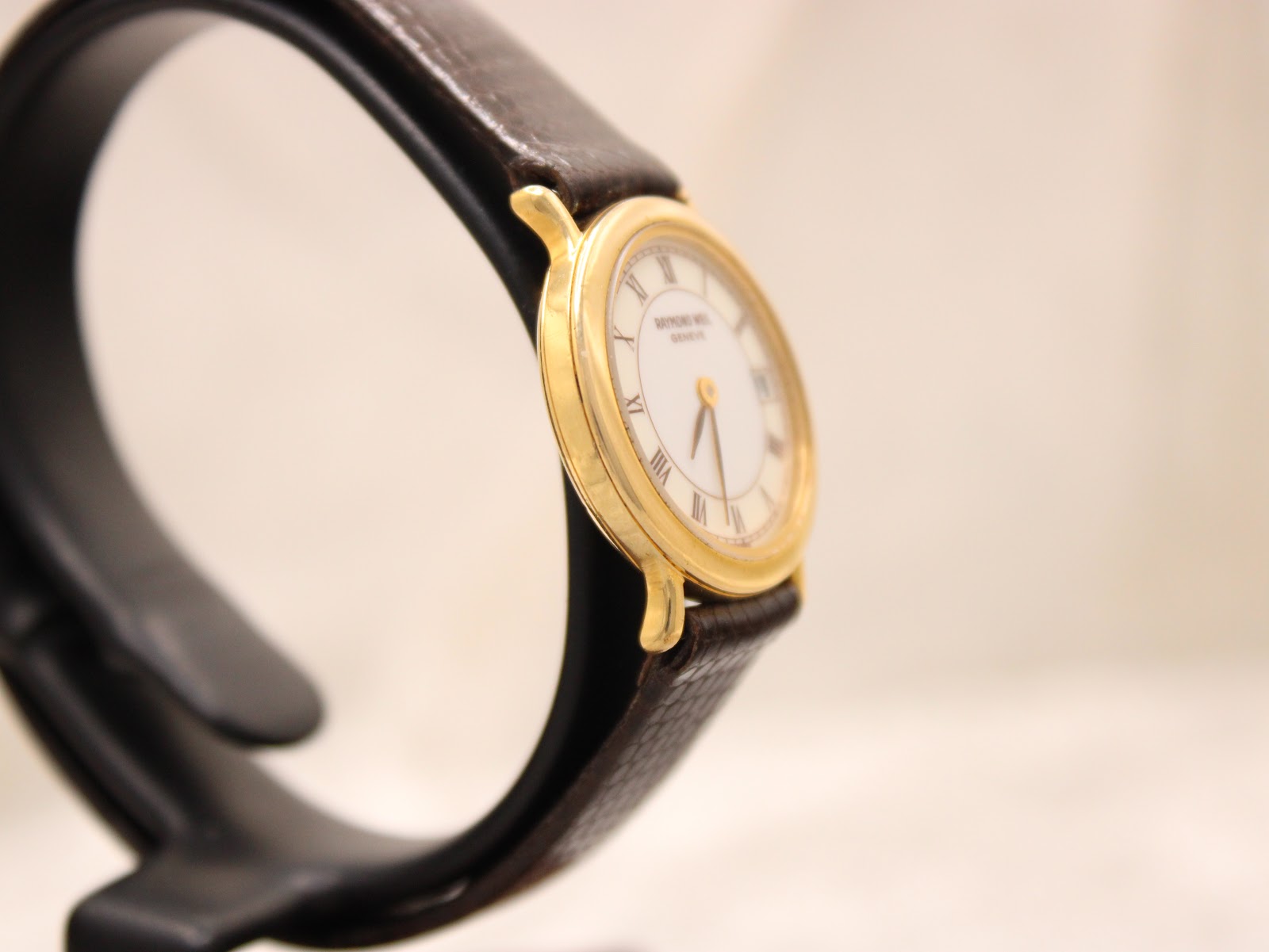 Angled side view of the Raymond Weil watch on a stand, showing the gold case and textured brown leather strap.