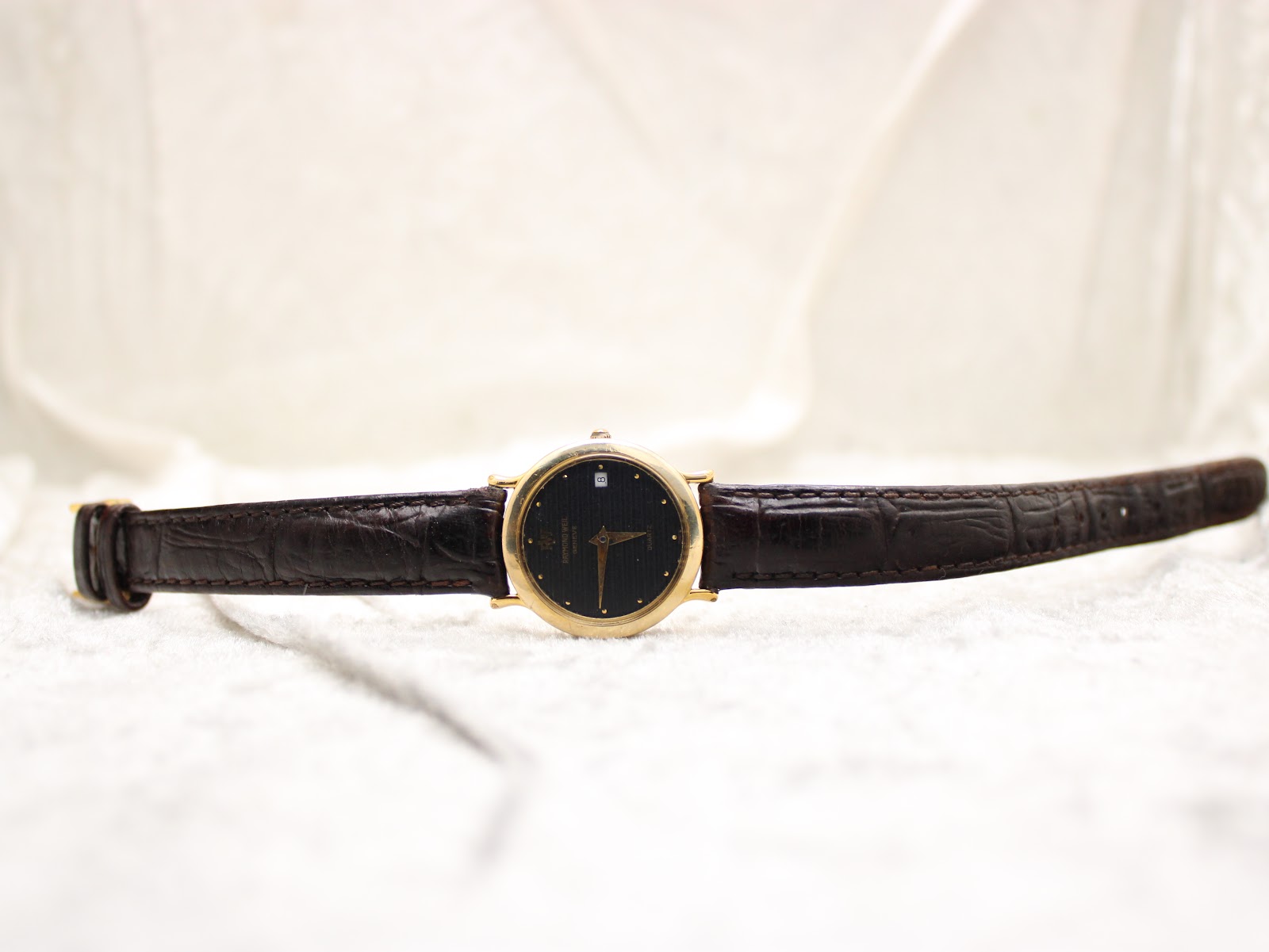 Full front view of the Raymond Weil watch with its black leather strap laid flat on a white textured surface.