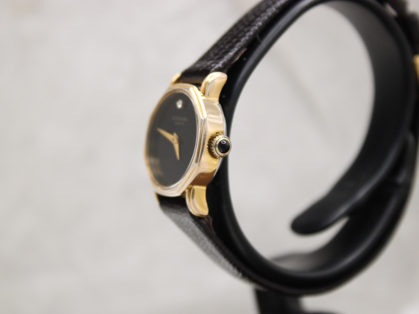Side profile view of the gold-tone Raymond Weil Geneve watch, showing the cabochon-set crown and textured black leather strap.