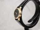 Side profile view of the gold-tone Raymond Weil Geneve watch, showing the cabochon-set crown and textured black leather strap.