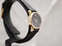 Angled side view of the Raymond Weil Geneve watch on a display stand, highlighting the slim gold-tone case and black strap.