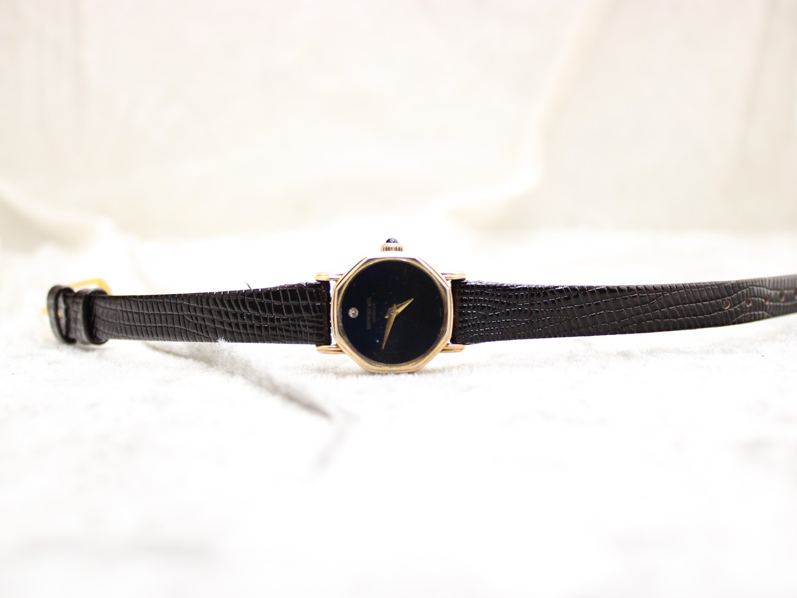 Full length top-down view of the Raymond Weil Geneve watch with its black leather strap laid flat on a white textured surface.