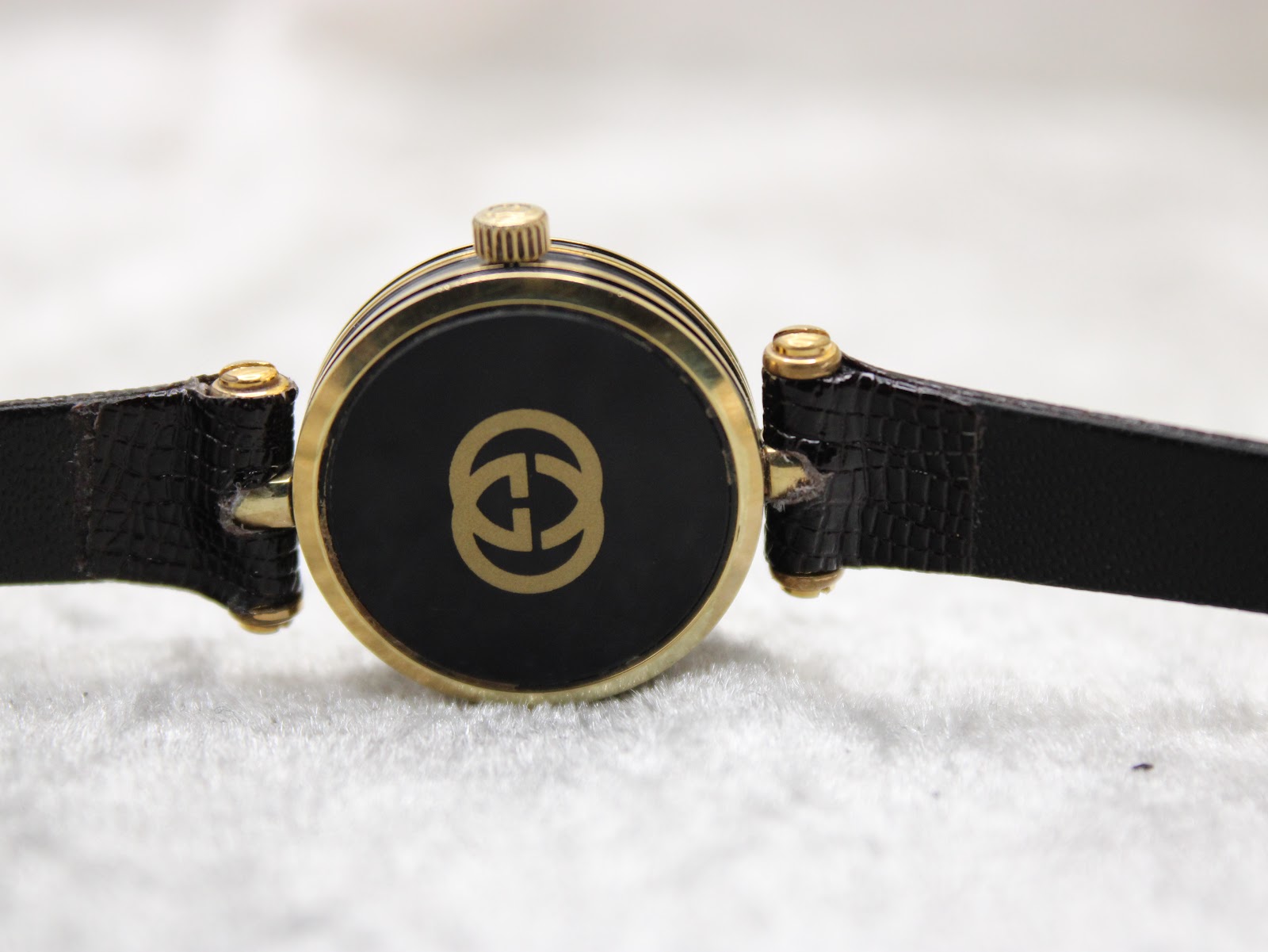 Close-up of the watch case back, showing the iconic Gucci interlocking G logo in gold against a black background.