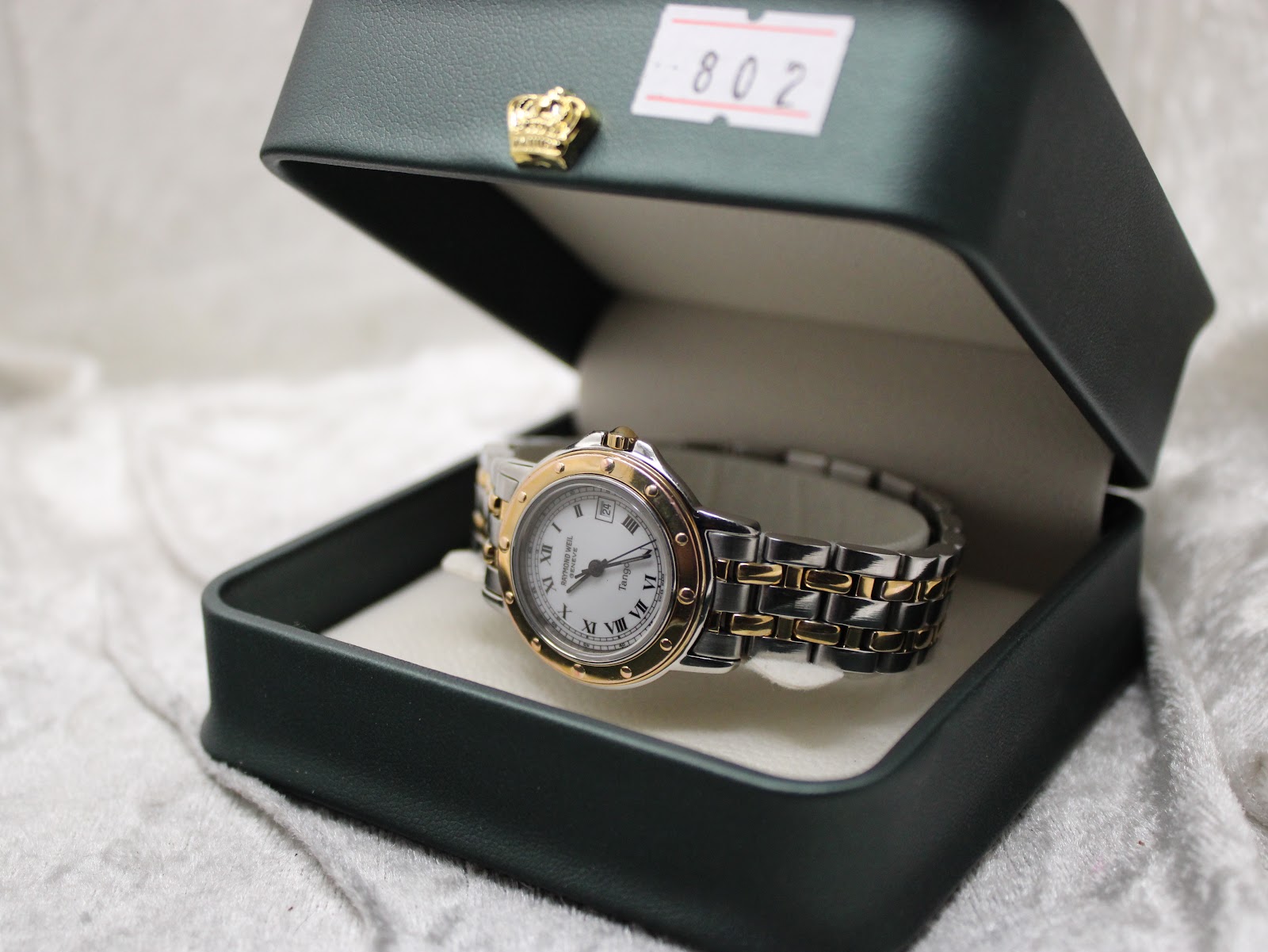 The Raymond Weil Tango watch resting inside a dark green presentation box, with a white label marked '802' visible in the background.