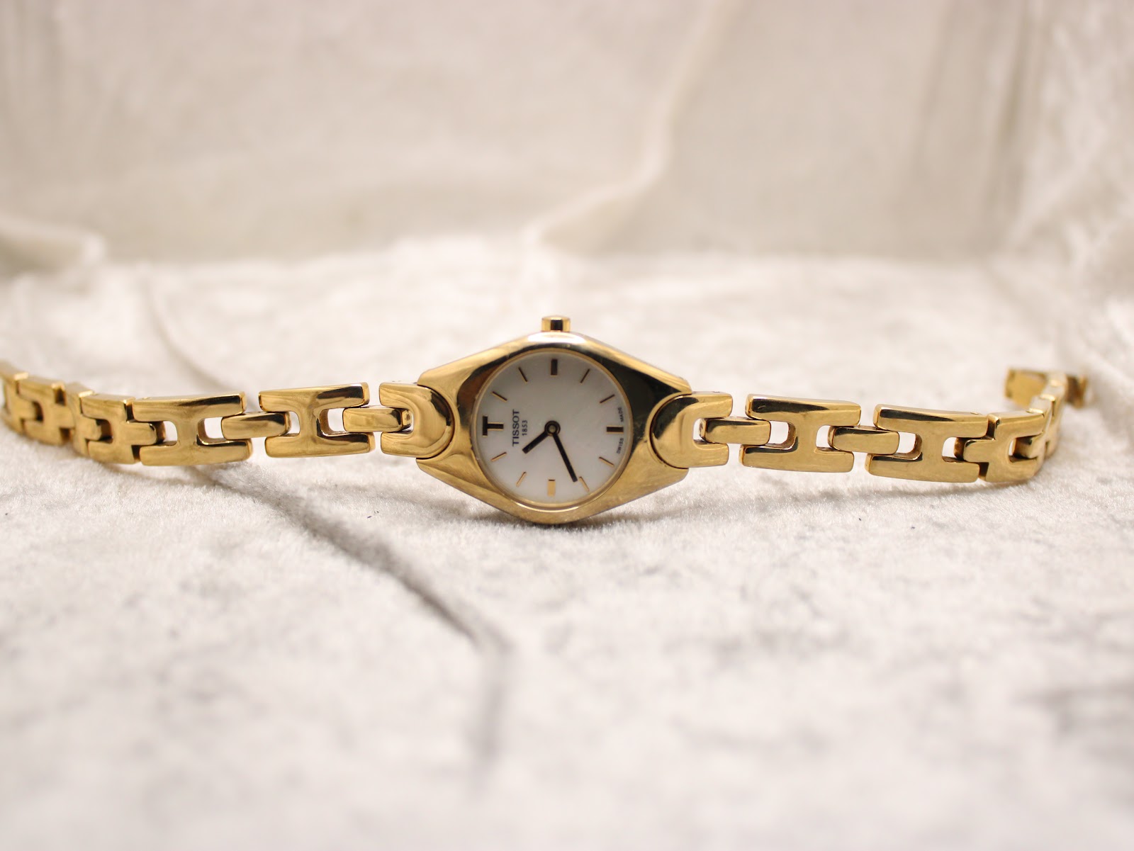 Top-down view of the gold-tone Tissot G349 watch with its bracelet fully extended on a white textured surface.
