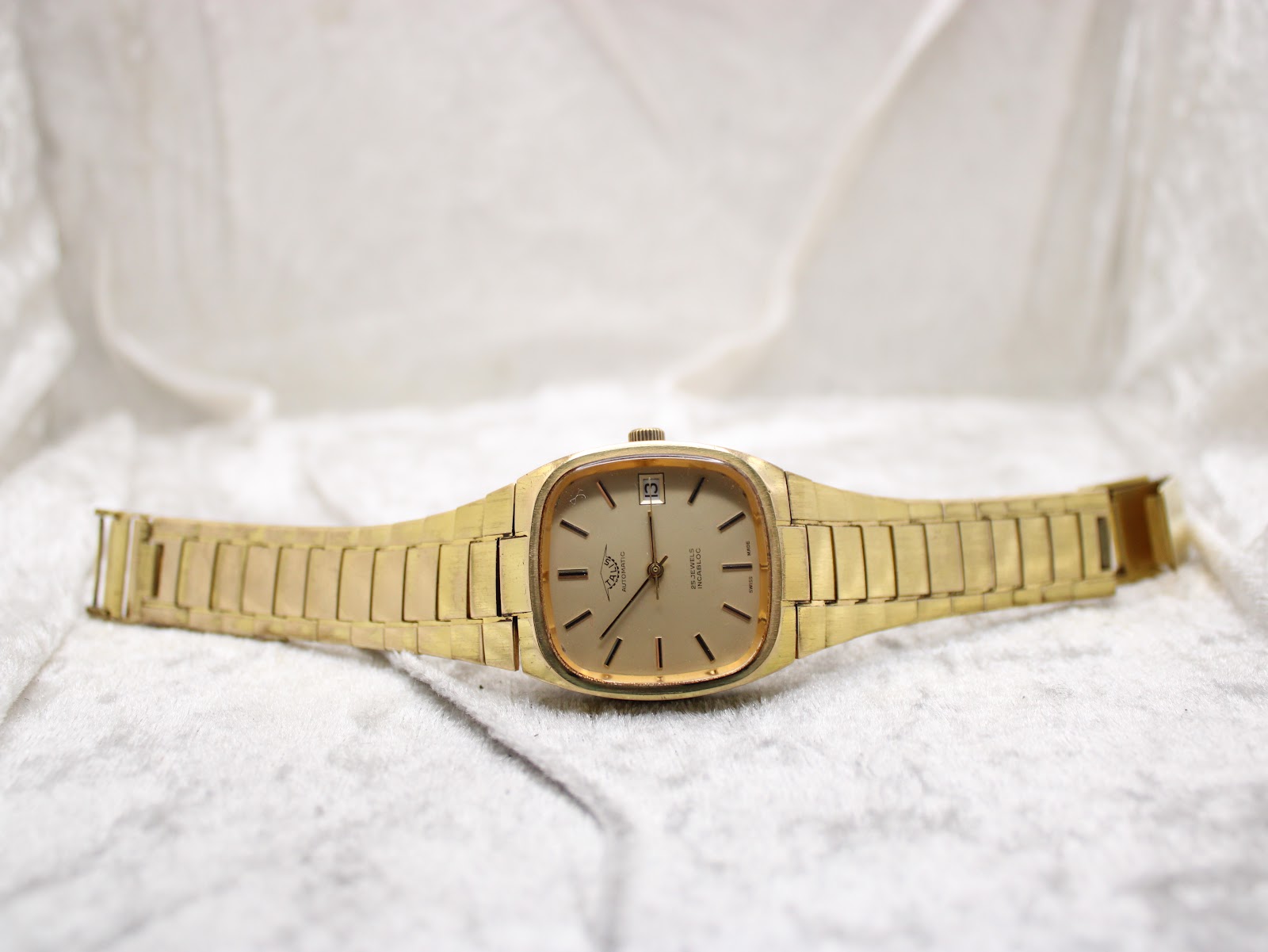 Full front view of the gold-tone Talis watch with its bracelet laid flat on a white textured surface.