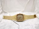Full front view of the gold-tone Talis watch with its bracelet laid flat on a white textured surface.