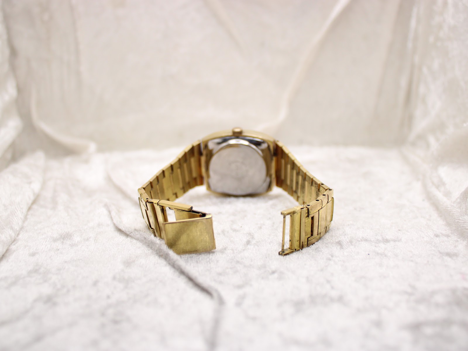 Back view of the entire gold-tone Talis watch and bracelet, showing the open folding clasp on a white textured surface.