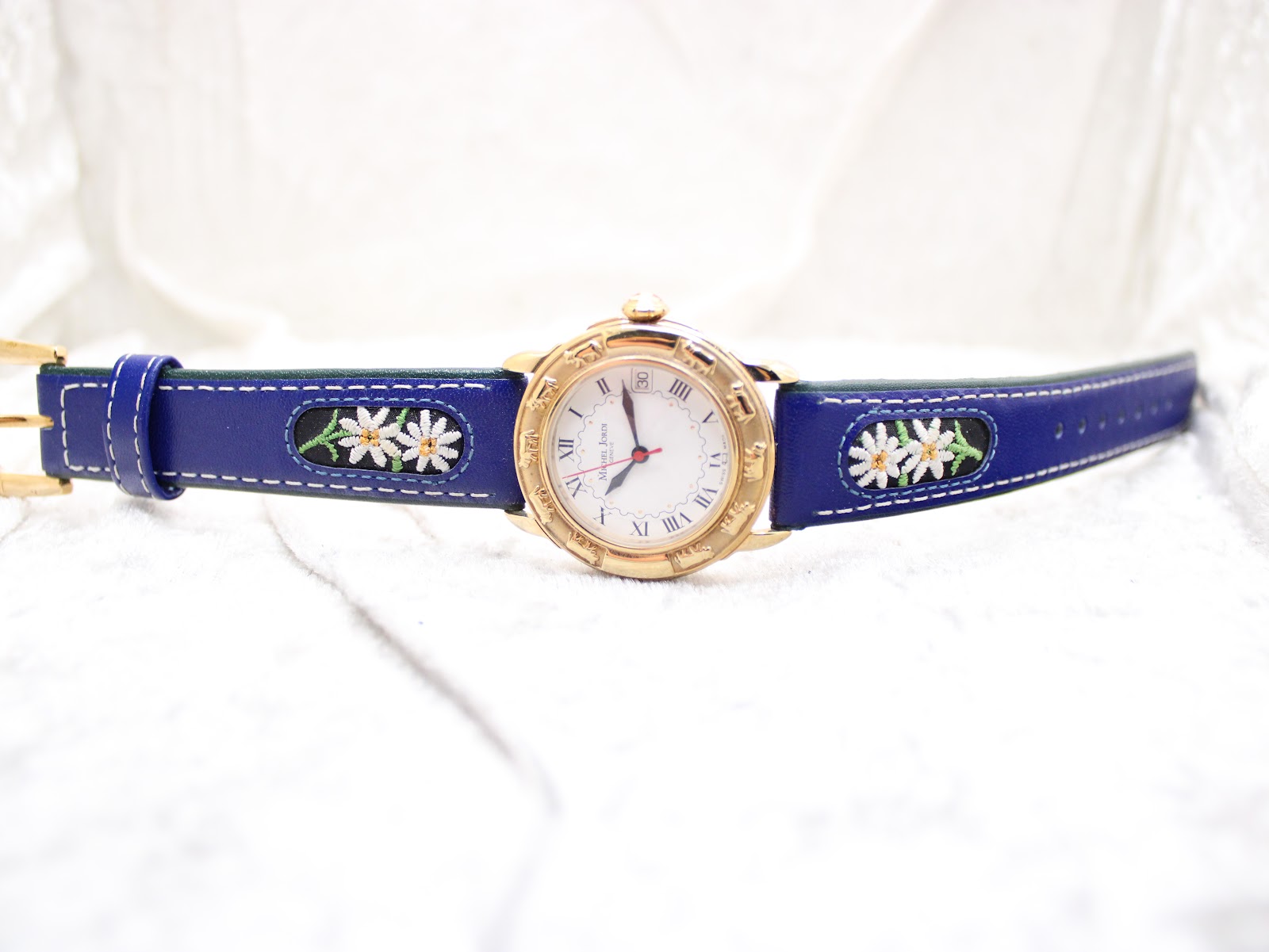 Full view of the Michel Jordi Genève watch and its blue embroidered leather strap laid flat on a white surface.