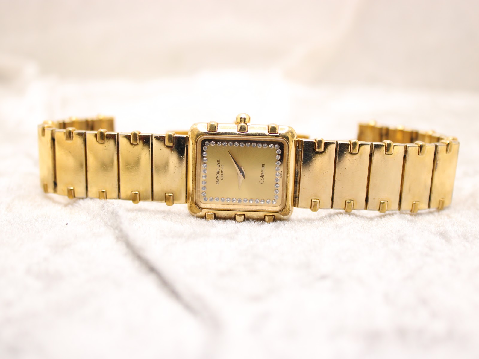 Full front view of the gold-plated Raymond Weil Coliseum watch with bracelet extended on a white textured surface.