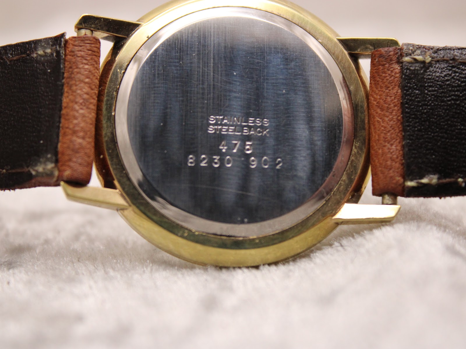 Close-up of the polished stainless steel case back with serial numbers "8230 902" and "475" engraved. Part of the light brown strap is visible.