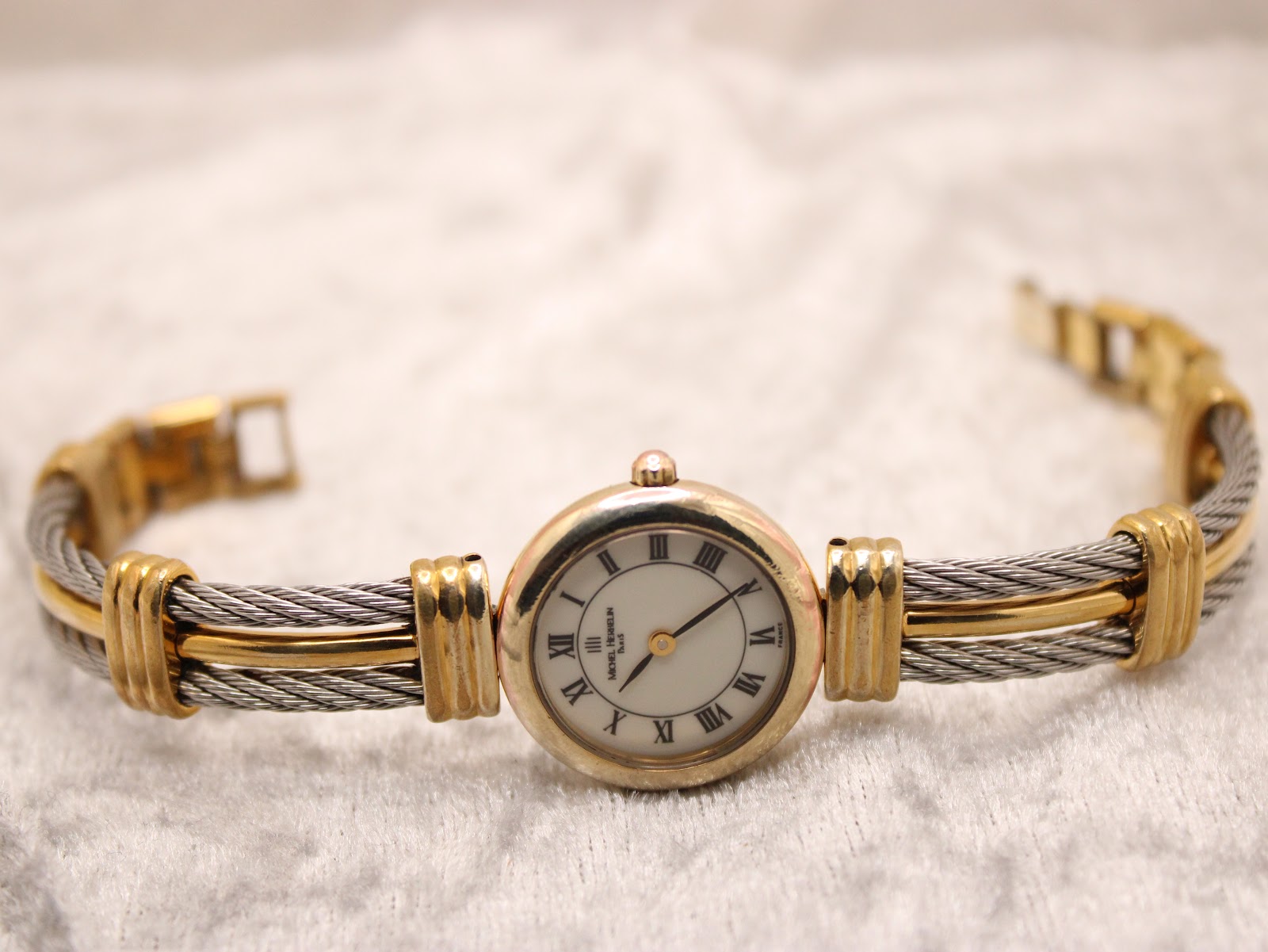 Full front view of the Michel Herbelin watch with its two-tone cable bracelet laid flat on a textured white surface.