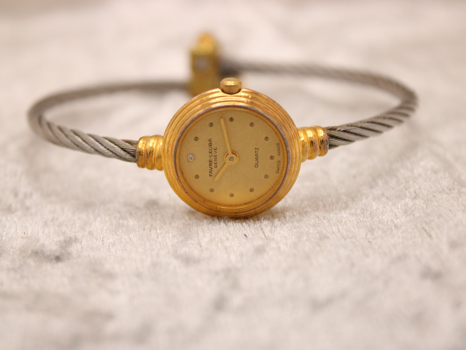 Top-down view of the watch on a soft surface, displaying the gold-tone dial with dot markers and the two-tone cable bracelet.