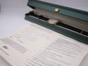 Overhead view of the watch inside a green presentation box, with the maintenance guarantee booklet showing a hand-filled service record in the foreground.