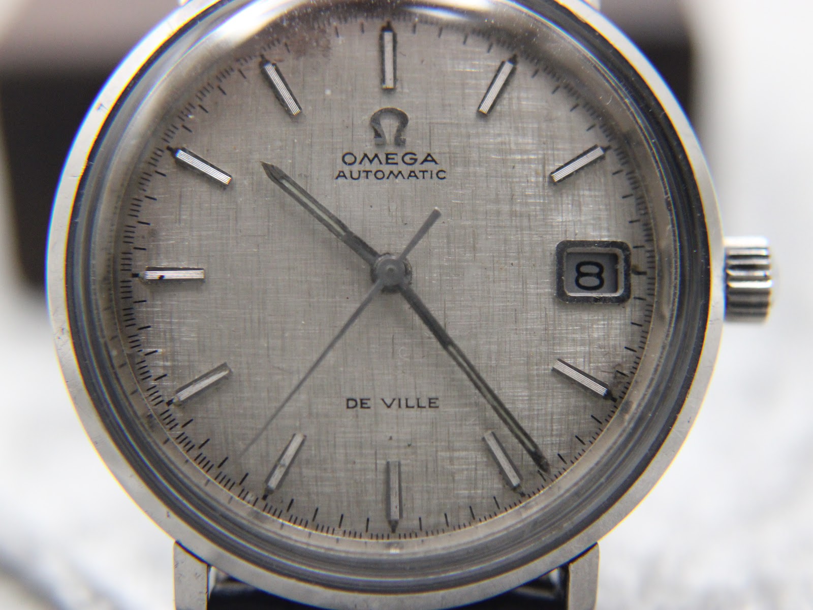 Close-up of the silver linen textured dial of the Omega De Ville, showing the Omega logo, "AUTOMATIC" and "DE VILLE" text, and the framed date window.