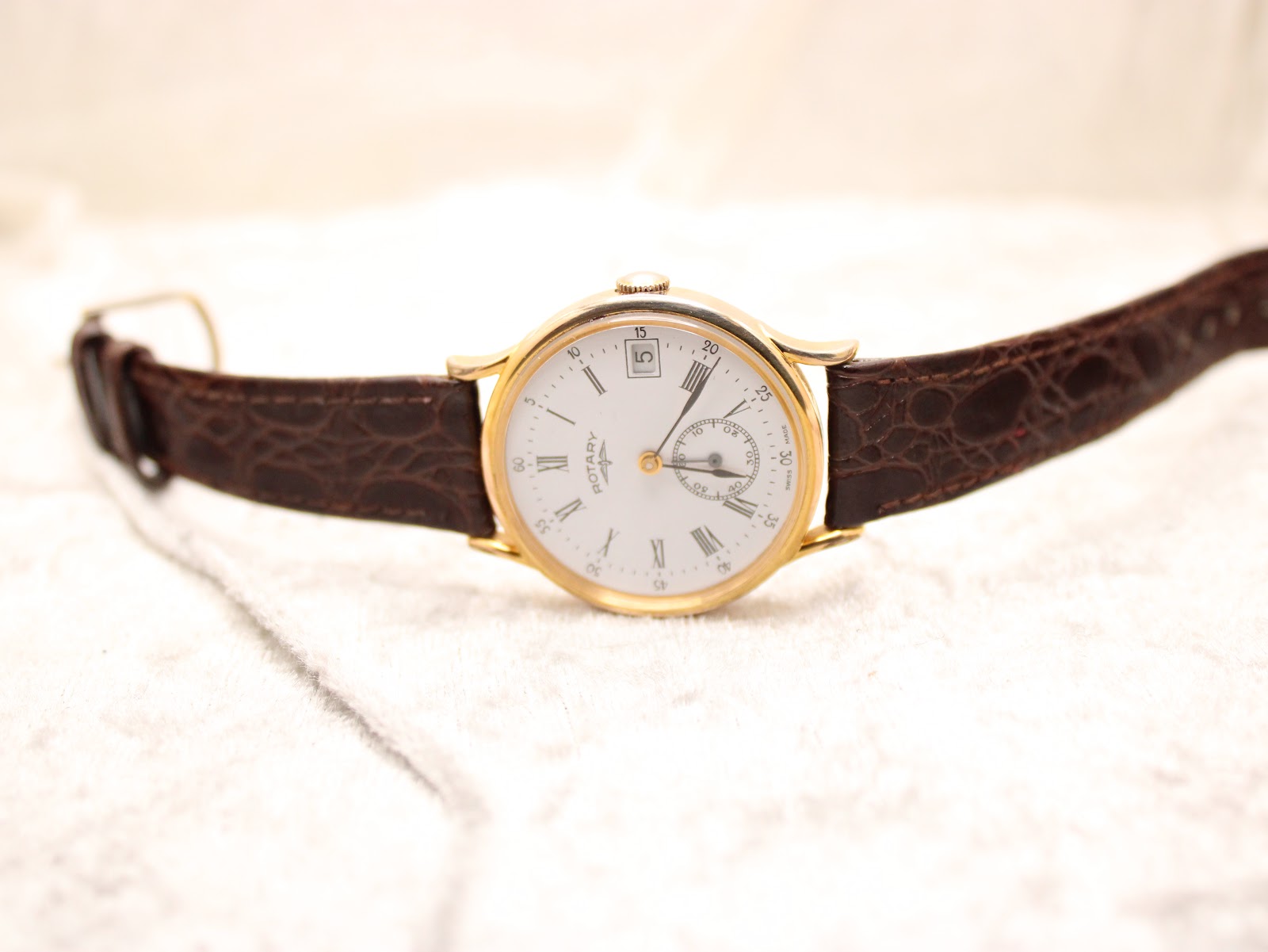 Overhead view of the Rotary watch with its brown leather strap laid flat on a white textured surface.
