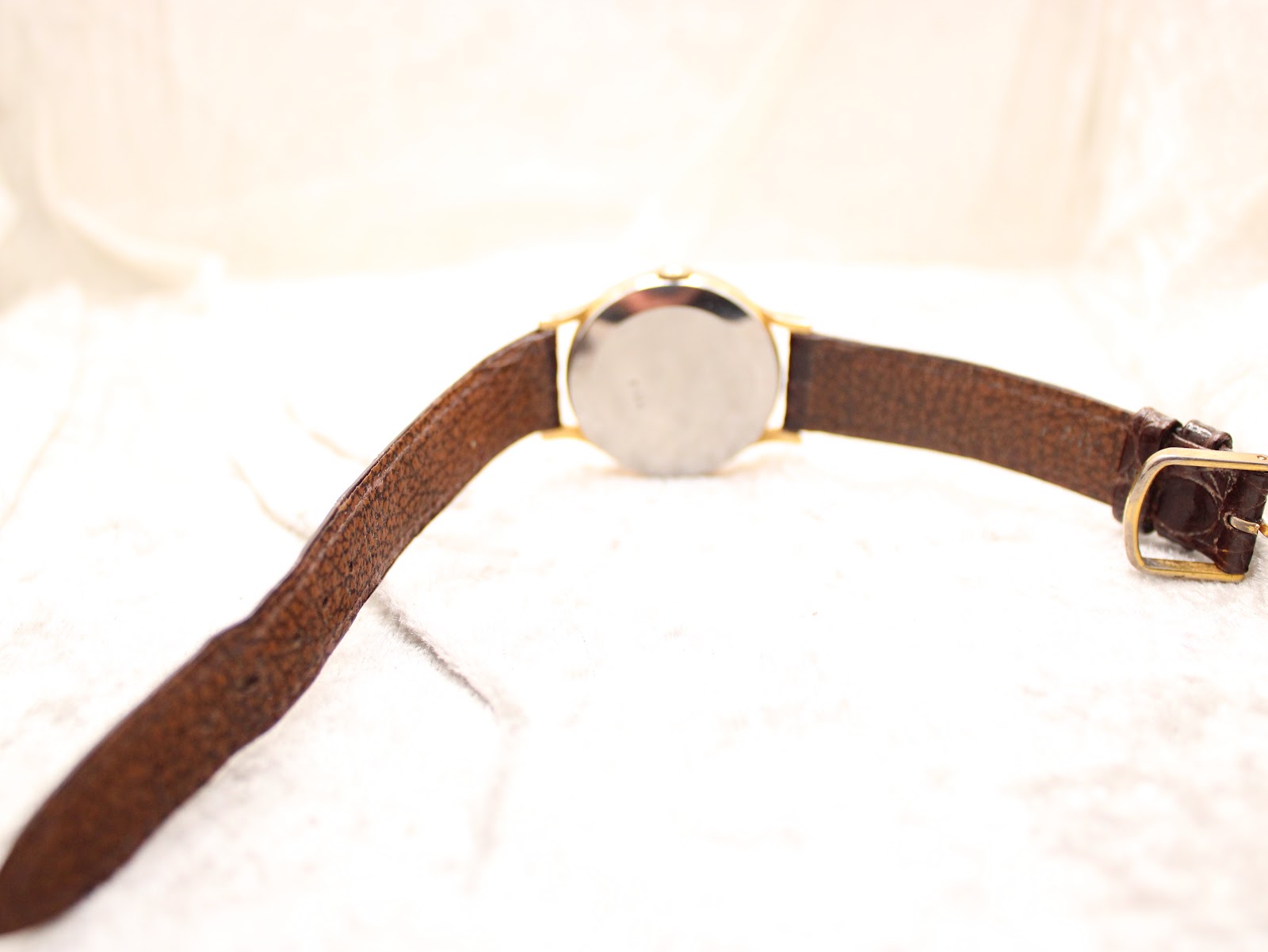 Rear view of the Rotary watch on a white textured surface, showing the plain stainless steel case back and brown strap.