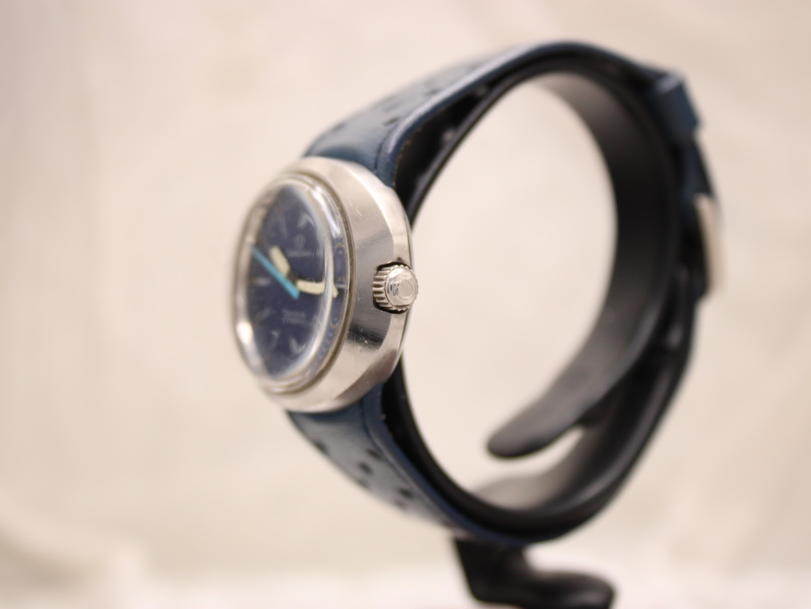 Side profile view of the Omega Genève Dynamic watch showing the signed crown and the curved case with a blue racing strap.